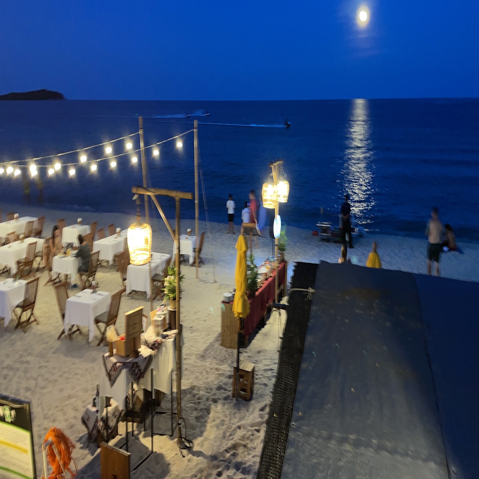 Beach restaurant tables on the sand under string lights and a full moon
